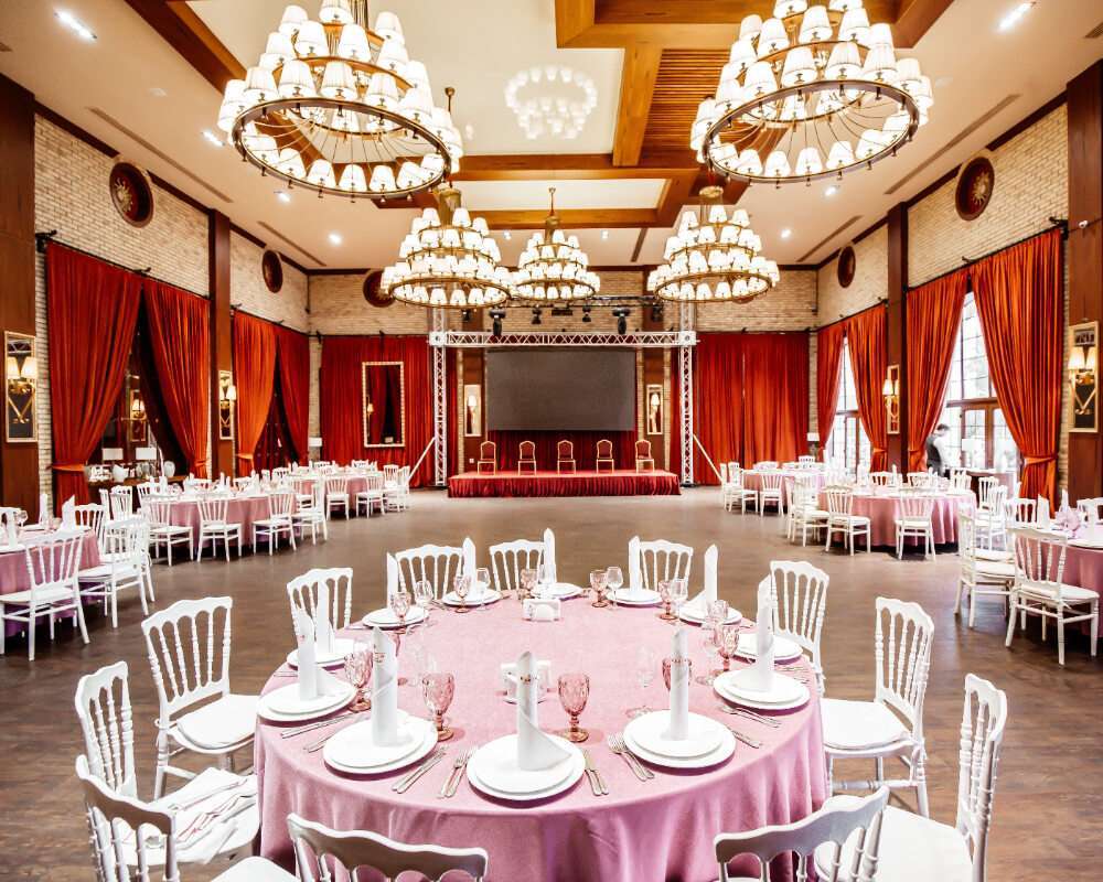 A luxurious indoor event space with elegant pink tablecloths, white chairs, and grand chandeliers, ideal for upscale event rentals and celebrations.