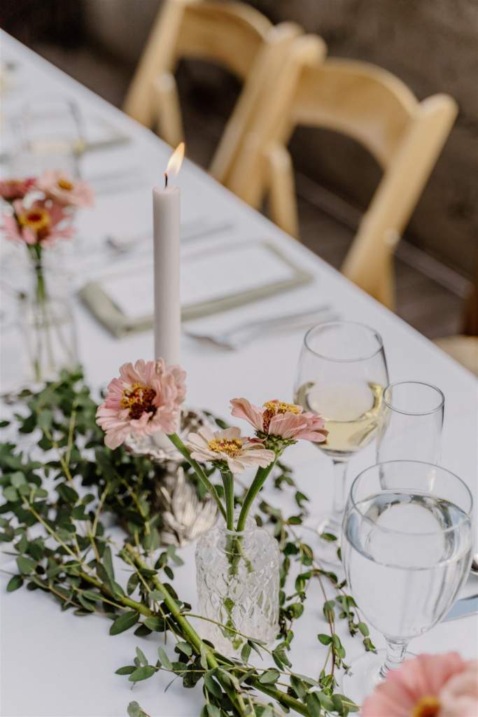 Elegant wedding table setting featuring a white tablecloth, pink floral centerpieces, a tall candle, wine glasses, and greenery along the table, with wooden chairs in the background.