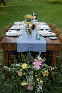 Outdoor dining setup with a long wooden table, blue table runner, floral centerpiece, wine glasses, and plates, surrounded by vibrant flower arrangements at the base, set against a green lawn.