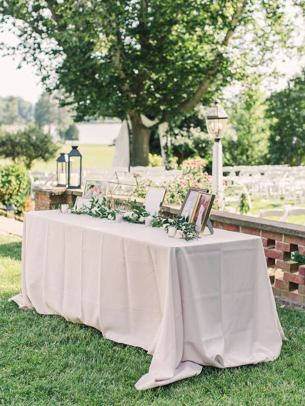 A beautifully arranged table outdoors with a simple floral display and framed pictures. It appears to be part of a wedding setup, likely using wedding rental items.