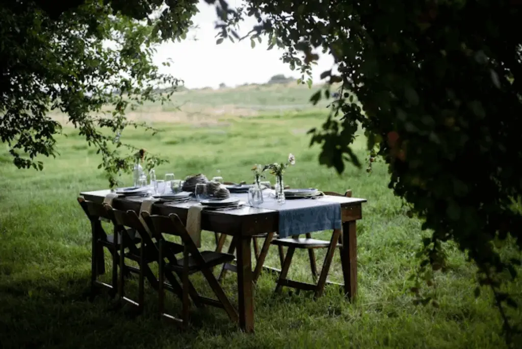 A beautifully set table for a small outdoor event, surrounded by trees, featuring elegant dinnerware and floral arrangements. This setup is ideal for small event planning rental.