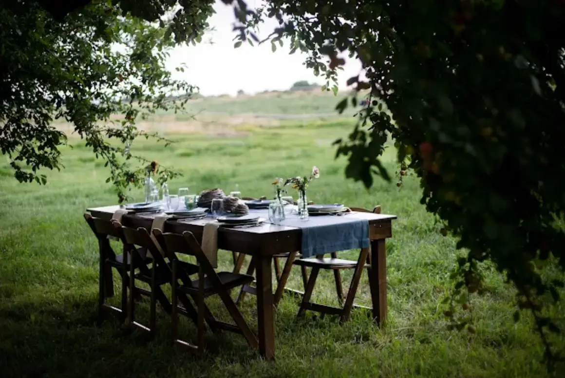 A beautifully set table for a small outdoor event, surrounded by trees, featuring elegant dinnerware and floral arrangements. This setup is ideal for small event planning rental.